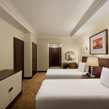 Hilton Suites Jabal Omar Makkah Mekka Exterior foto The photo shows a hotel room that features two beds with white linens. The room has light-colored walls and a warm-colored carpet. There is a wooden dresser with a flat-screen TV mounted on the wall opposite the beds. To the side, there are two doors