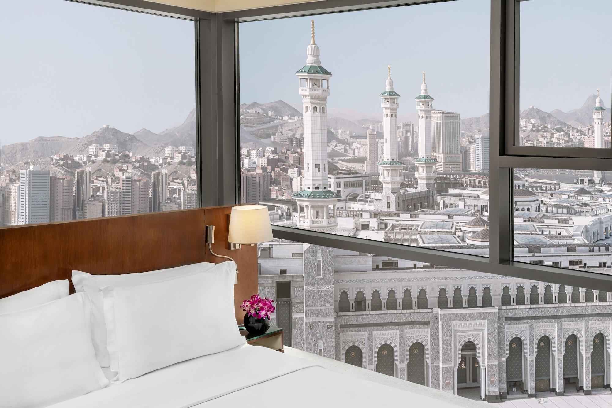 Hilton Suites Jabal Omar Makkah Mekka Exterior foto The photo depicts a hotel room with a modern design. In the foreground, there is a neatly made bed with white linens and a bedside lamp. A small flower arrangement adds a touch of color to the room. Through large windows, a stunning view of a citysca