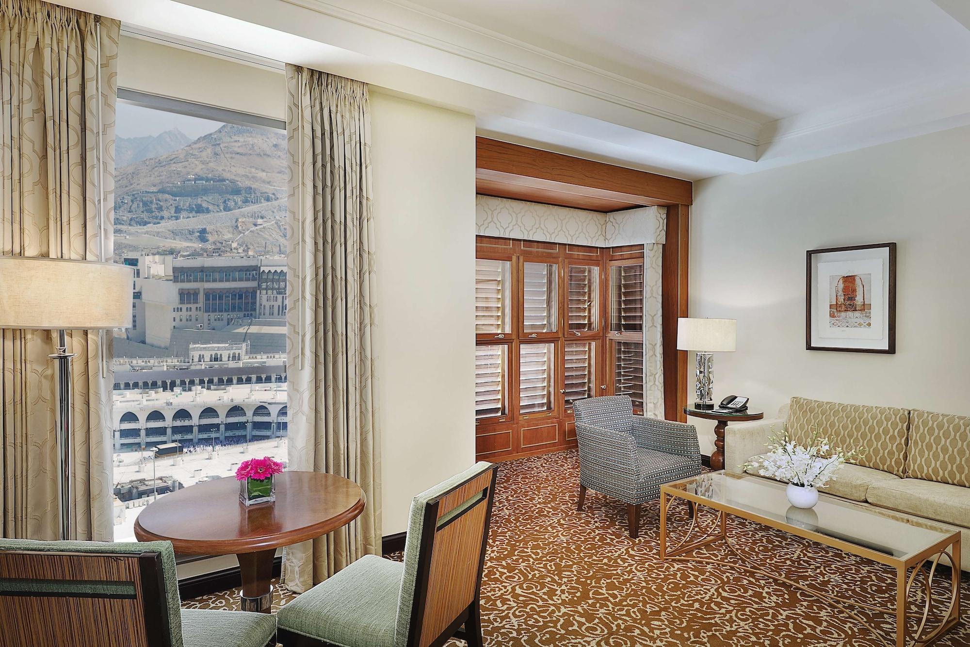Hilton Suites Jabal Omar Makkah Mekka Exterior foto The photo shows a luxurious hotel room with a welcoming ambiance. There are large windows that provide a view of the outside, suggesting a well-lit space. The room features a seating area with a couch and chairs, alongside a small round dining table.