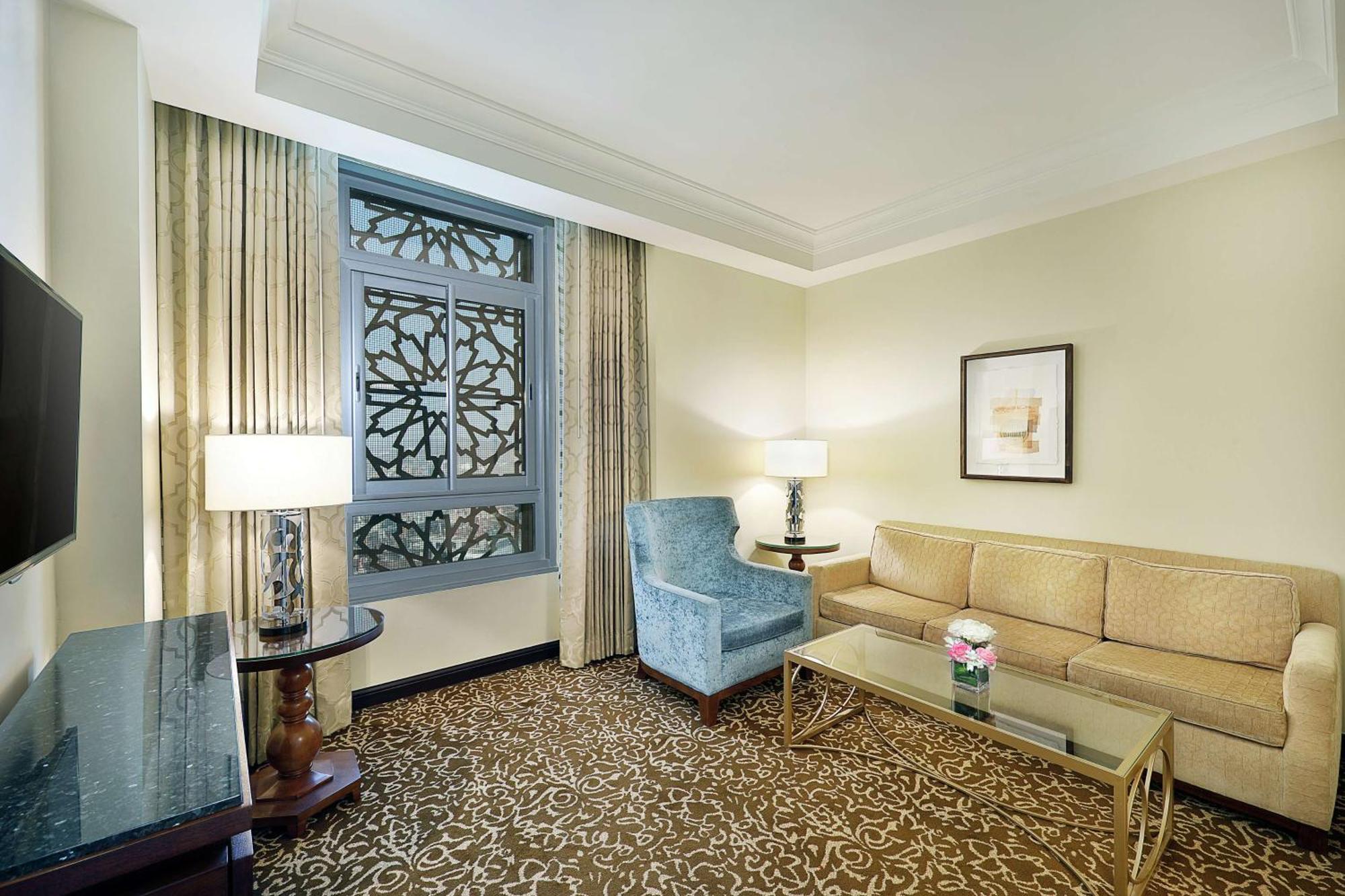 Hilton Suites Jabal Omar Makkah Mekka Exterior foto The photo shows a well-decorated hotel room or suite. It features a light-colored wall with a decorative window that has an intricate design. There are two windows adorned with curtains. The furniture includes a beige couch, a blue upholstered armcha