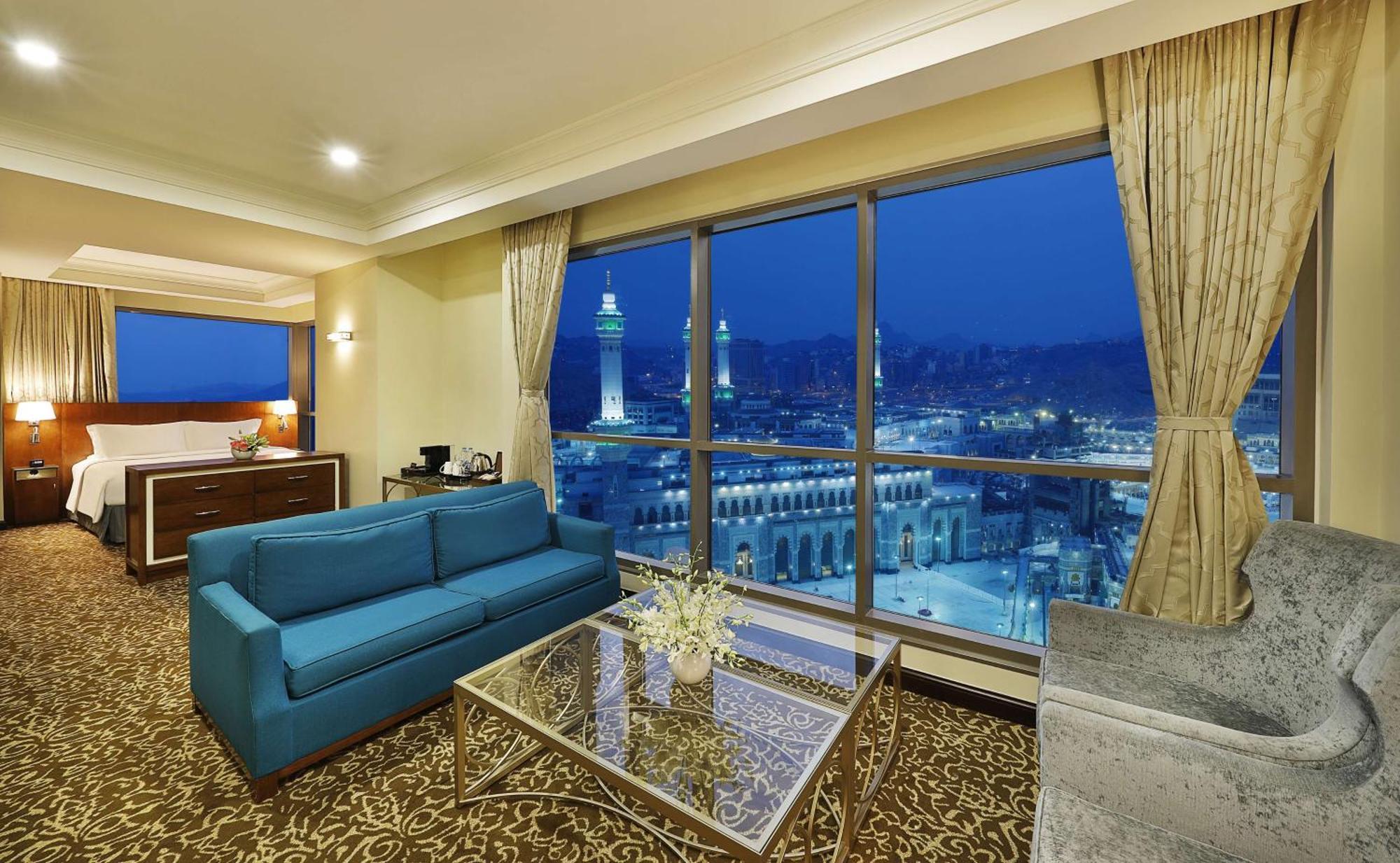 Hilton Suites Jabal Omar Makkah Mekka Exterior foto The photo shows a luxurious hotel room with a stunning view. The room features a modern design, including a blue sofa and a glass coffee table on a patterned carpet. Large windows provide an expansive view of a cityscape, illuminated at night, showca