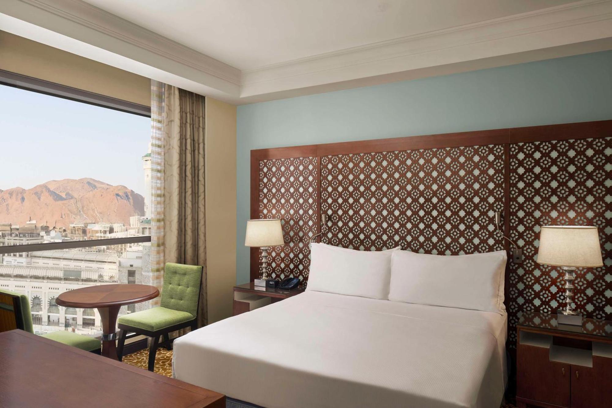 Hilton Suites Jabal Omar Makkah Mekka Exterior foto The photo shows a hotel room featuring a large bed with white linens and a decorative headboard. Beside the bed, there are two lamps on nightstands. A small round table with a green chair is positioned near the window, which offers a view of a mounta