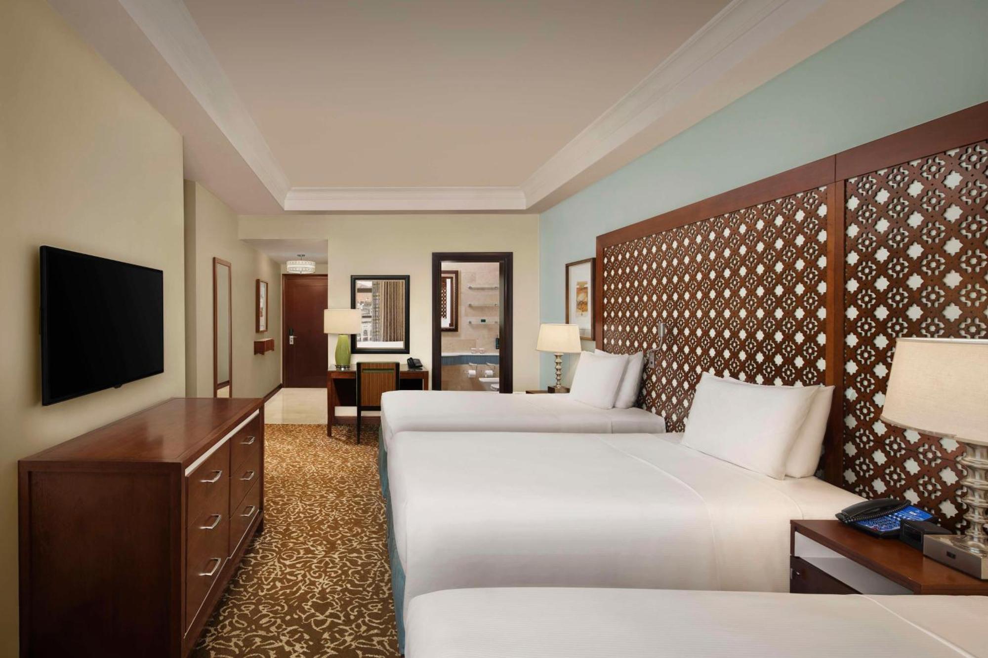 Hilton Suites Jabal Omar Makkah Mekka Exterior foto The photo shows a hotel room featuring two beds with white linens. There is a decorative headboard behind each bed, made of wood with a patterned design. The room has a warm beige carpet and light blue walls, giving it a serene atmosphere. A wooden d