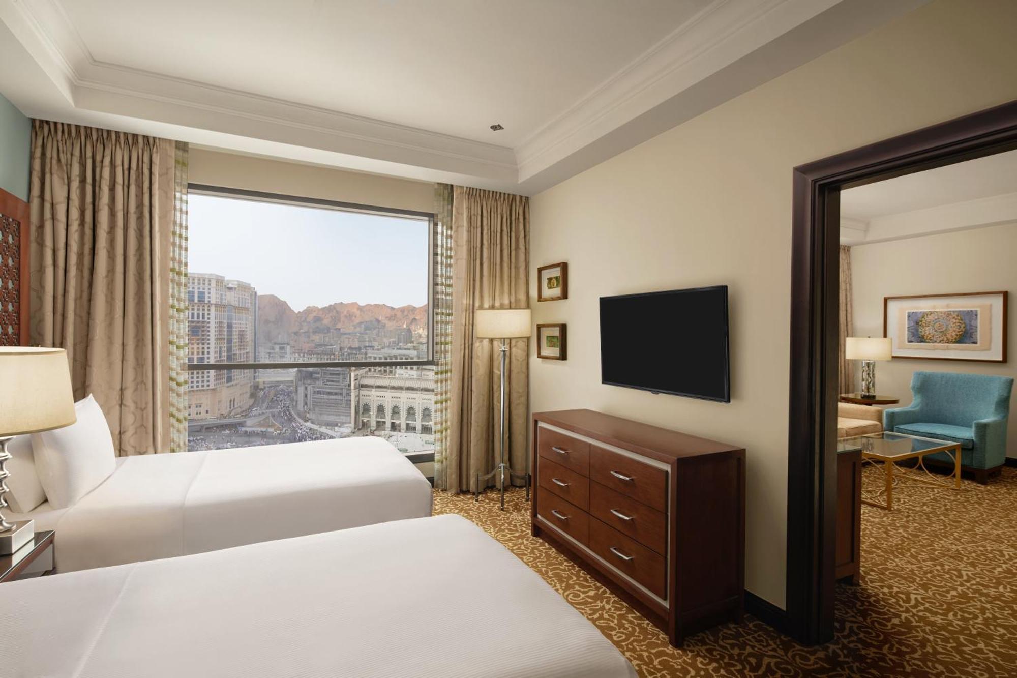 Hilton Suites Jabal Omar Makkah Mekka Exterior foto The photo shows a hotel room featuring two beds with white bedding. There is a large window with curtains that allow for natural light, offering a view of the outside scenery, which includes mountains. In the room, there is a wooden dresser and a fla
