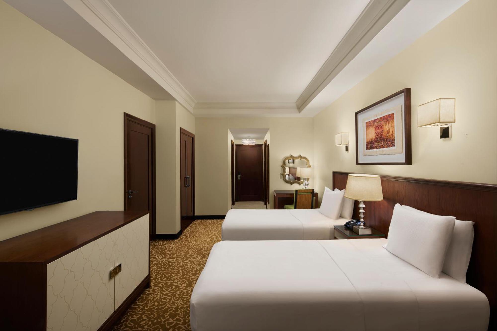 Hilton Suites Jabal Omar Makkah Mekka Exterior foto The photo shows a hotel room that features two beds with white linens. The room has light-colored walls and a warm-colored carpet. There is a wooden dresser with a flat-screen TV mounted on the wall opposite the beds. To the side, there are two doors