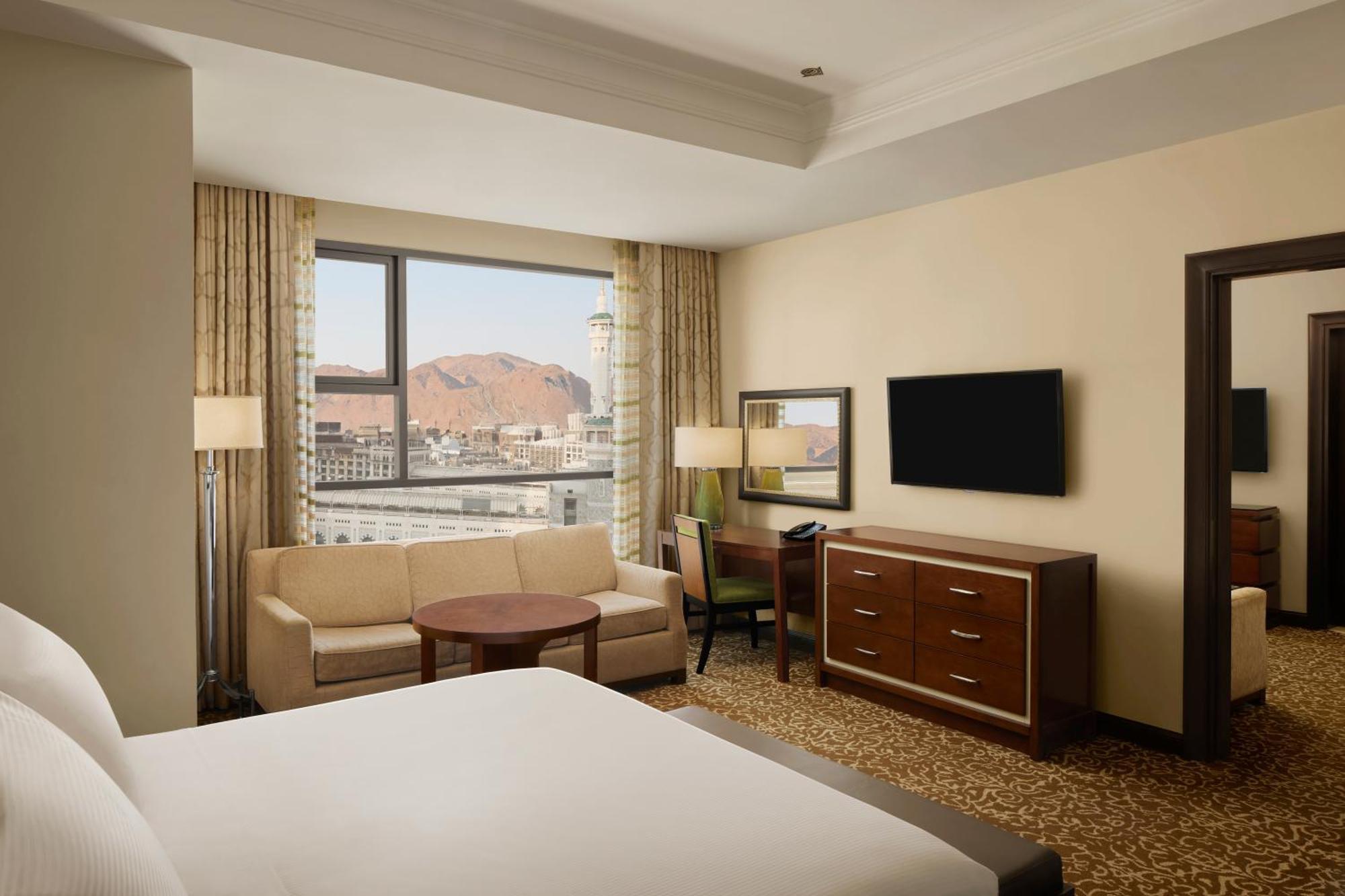 Hilton Suites Jabal Omar Makkah Mekka Exterior foto The photo shows a hotel room with modern decor. In the foreground, there is a large bed with white linens. To the left, a comfortable seating area features a sofa, a round coffee table, and a chair. A large window offers a view, likely of a mountain 